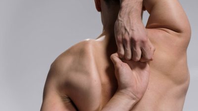 Shirtless muscular man practicing a yoga stretch with hands clasped behind back on a white background.