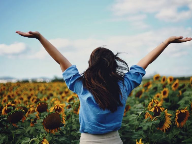 A woman in a blue shirt joyfully raises her arms amidst a sunflower field in Izmir, Türkiye.